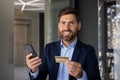 Smiling and successful young man businessman and banker standing in office, holding phone and credit card, looking Royalty Free Stock Photo