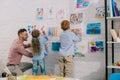 smiling teacher helping little preschoolers hang colorful pictures on wall