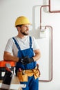 Smiling technician, handyman in uniform, hard hat and protective gloves looking away while standing indoors, leaning on Royalty Free Stock Photo