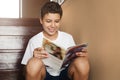 Smiling teen boy sitting at home on steps, reading magazine. Cheerful guy in white t-shirt is sitting at home, reading comic book