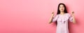 Smiling teen girl asian pointing fingers up at empty space, advertising on pink background