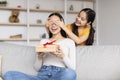 Smiling teen girl congratulates surprised young asian lady, gives box with present, closes eyes to mom