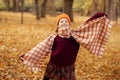 Smiling teenage girl stretching hands, holding checkered scarf, standing on yellow fallen maple leaves in park forest.