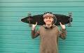 Smiling teenager in a cap and a hoodie stands on the background of a green wall, holds a longboard on his shoulde