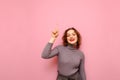 Smiling teenager girl with curly hair and in a gray sweater stands on a pink background and shows her index finger up at a blank Royalty Free Stock Photo