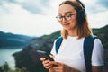 Smiling traveler woman with hipser glasses and backpack holds smartphone device in hands and listens music for headphones Royalty Free Stock Photo