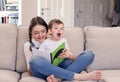 Smiling tween girl reading book to her cute little brother sitting on her lap and yawning on sofa at home. Siblings care and love.