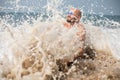 Smiling vacationist in sunglasses covered by wave sitting on sandy seaside. Splash of water scattered on foreground.