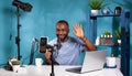 Smiling vlogger waving hello in front of recording smartphone sitting at desk