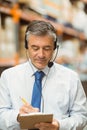 Mature man wearing headset and blue necktie, checking clipboard and writing with pencil by racking Royalty Free Stock Photo