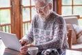 Smiling white haired senior man in winter sweater sitting in living room using laptop computer. Carefree elderly grandfather Royalty Free Stock Photo