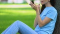 Smiling woman applying lip gloss using hand mirror, sitting on grass in park