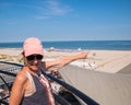 Smiling woman baby boomer dressed casually with pink hat on balcony pointing to the beach and ocean Royalty Free Stock Photo