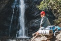 Smiling woman with backpack in red hat dressed in active trekking clothes and boots sitting near mountain river waterfall and Royalty Free Stock Photo