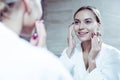 Beautiful young woman smiling while looking into mirror and using cream Royalty Free Stock Photo