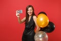 Smiling woman in black dress celebrating holding air balloons doing taking selfie shot on mobile phone isolated on red Royalty Free Stock Photo