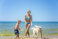 Smiling woman in a blue swimsuit with a little boy in swimming trunks and a white dog on a sandy beach at the sea on a sunny day. Royalty Free Stock Photo