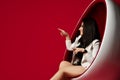 Smiling woman brunette in white long jacket sitting in a modern oval chair and pointing her finger at something on red Royalty Free Stock Photo