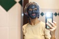 Smiling woman with clay mask taking a photograph with mobile phone