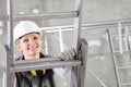 Smiling woman construction worker builder on ladder wearing white helmet and hearing protection headphones on interior site