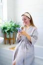 Smiling woman with facial pink clay mask on the face using smartphone at home bath enjoying relaxation and spa beauty Royalty Free Stock Photo