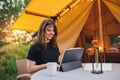 Smiling Woman freelancer using a laptop on a cozy glamping tent in a sunny day. Luxury camping tent for outdoor summer holiday and Royalty Free Stock Photo