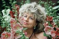 Smiling woman in her beautiful garden. A Mature woman stands among beautiful tall pink hollyhocks