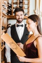 smiling woman holding menu card at wine store while steward standing blurred