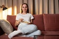 Smiling woman at home sitting on the couch and watching tv, she is holding a remote control and eating popcorn Royalty Free Stock Photo