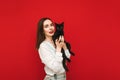 Smiling woman in light casual clothes stands on a red background with a black cat in her hands, looks into the camera and smiles. Royalty Free Stock Photo