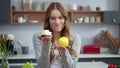 Smiling woman looking at cake and apple in kitchen. Girl preferring fresh fruit.