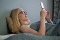 Smiling woman looking at mobile phone lying down on white bed. Happy blond young woman using cellphone at home. Royalty Free Stock Photo