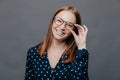 Smiling woman with pleased facial expression, keeps hand on rim of spectacles, wears black polka dot blouse, rejoices promotion at