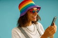 Smiling woman in rainbow cap is looking on mobile phone standing over blue studio background Royalty Free Stock Photo