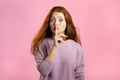 Smiling woman with red hair holding finger on her lips over pink background. Gesture of shhh, secret, silence. Close up. Royalty Free Stock Photo