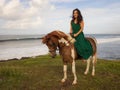 Smiling woman riding horse near the ocean. Outdoor activities. Asia woman wearing long green dress. Traveling concept. Cloudy sky Royalty Free Stock Photo