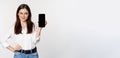 Smiling woman showing mobile phone screen, looking cunning, standing over white background