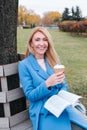 Smiling woman sits near a tree in an autumn park and holds a book and a cup with a hot drink in her hands. Royalty Free Stock Photo