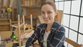 A smiling woman sits in a woodworking workshop surrounded by tools, embodying craftsmanship in a creative studio setting Royalty Free Stock Photo