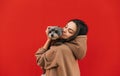 Smiling woman standing on a background of a red wall with a little cute dog in her arms, hugging a pet. Lady carries a pet in her Royalty Free Stock Photo