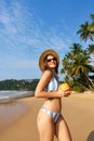 Smiling woman in striped bikini holds fresh coconut on sunny tropical beach, palm trees sway, clear sky, wellness travel Royalty Free Stock Photo