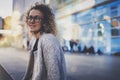 Smiling woman in stylish clothing wearing eye glasses outside in the european night city. Bokeh and flares effect on