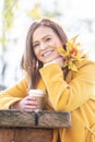 Smiling woman on a sunny autumn day outside holds a cup of coffee and couple of yellow leaves Royalty Free Stock Photo