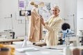 Smiling woman tailor shows new beige jacket shooting video while African-American seamstress sits at workplace