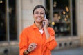 Woman talking by phone wearing orange shirt standing outdoors near building. Royalty Free Stock Photo
