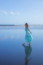 Smiling woman walking barefoot on empty beach. Full body portrait. Slim Caucasian woman wearing long dress. Water reflection.