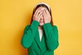 Smiling woman wearing green jacket posing isolated over yellow background hiding her eyes covering face with palms positive Royalty Free Stock Photo