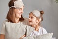 Smiling woman wearing sleeping mask sitting at home with her cute little daughter on bed talking looking at each other expressing Royalty Free Stock Photo