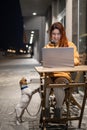 Smiling woman working on laptop at a wooden table in the street. The girl looks at the monitor and Jack Russell Terrier