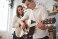 Smiling woman enjoys the performance. Young guitarist playing love song for his girlfriend in the kitchen Royalty Free Stock Photo